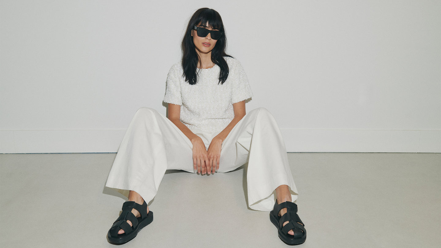 Person wearing a white outfit with black sandals sitting on a light gray floor.