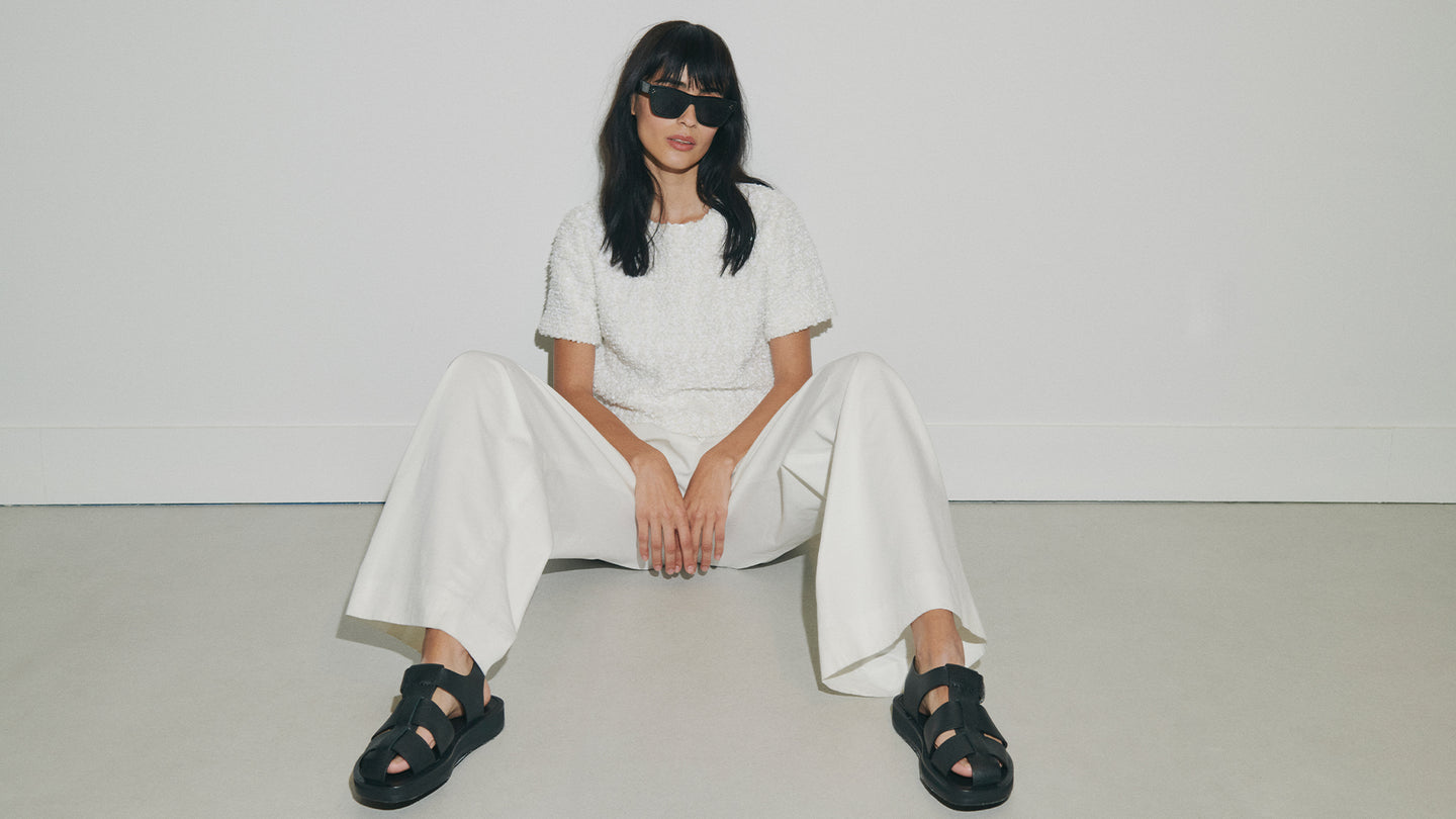 Person wearing a white outfit with black sandals sitting on a light gray floor.