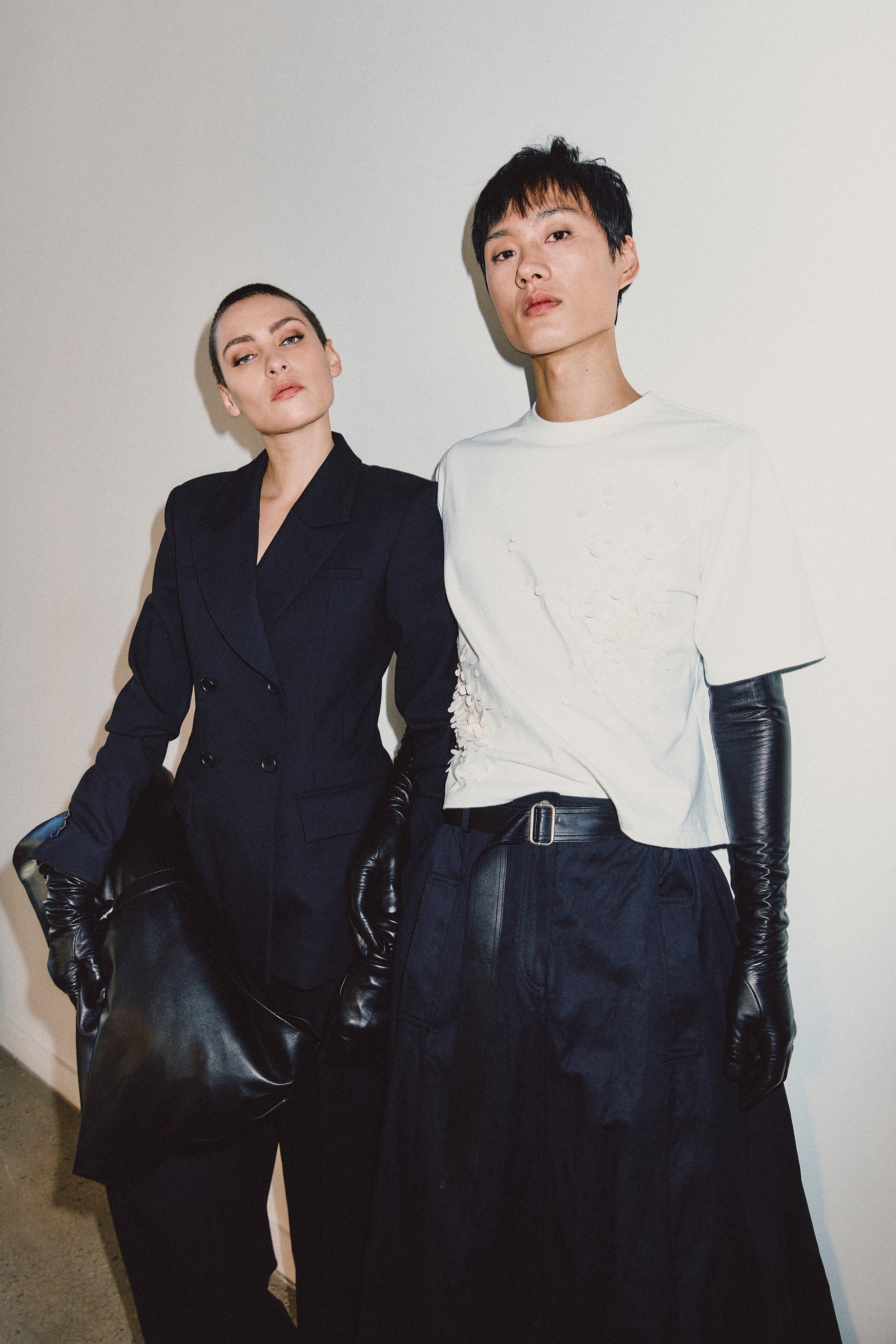 A fashion editorial photo featuring a model in a dark navy double-breasted blazer with black leather gloves and a matching model in a white graphic t-shirt, black leather gloves, and a long dark skirt, posed against a plain off-white wall.