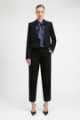 A model stands against a light gray studio background wearing a black cropped blazer and matching black trousers, styled with a blue patterned bow tie and black pointed heels.