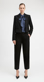 A model stands against a light gray studio background wearing a black cropped blazer and matching black trousers, styled with a blue patterned bow tie and black pointed heels.