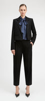 A model stands against a light gray studio background wearing a black cropped blazer and matching black trousers, styled with a blue patterned bow tie and black pointed heels.