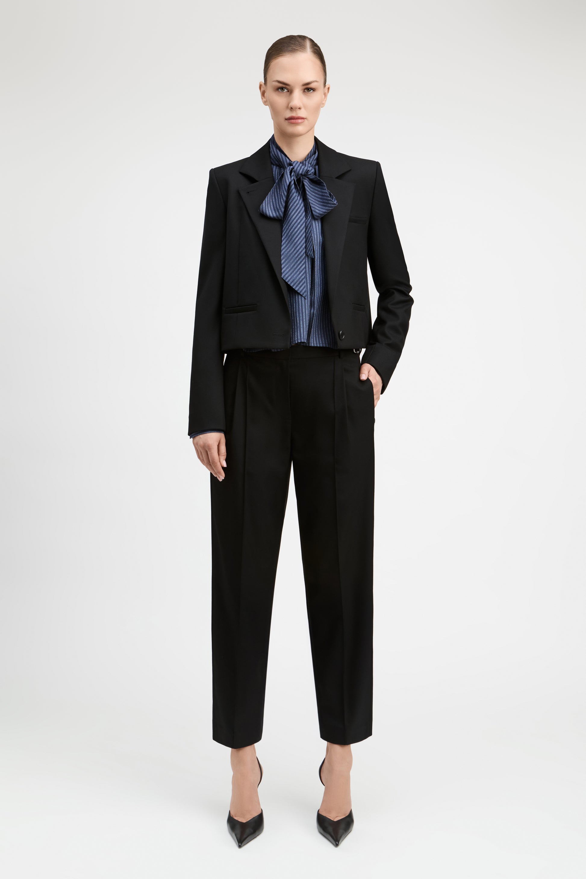 A model stands against a light gray studio background wearing a black cropped blazer and matching black trousers, styled with a blue patterned bow tie and black pointed heels.