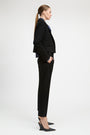 A woman in a black tailored blazer and matching cropped trousers, styled with a dark blue collared shirt and black slingback heels, shown in profile against a light gray studio background.