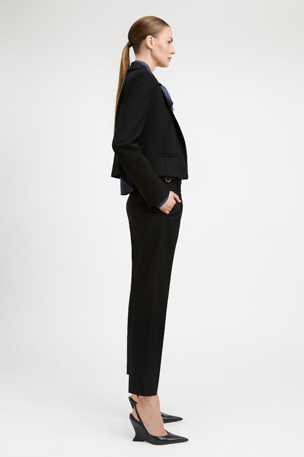 A woman in a black tailored blazer and matching cropped trousers, styled with a dark blue collared shirt and black slingback heels, shown in profile against a light gray studio background.
