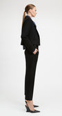 A woman in a black tailored blazer and matching cropped trousers, styled with a dark blue collared shirt and black slingback heels, shown in profile against a light gray studio background.