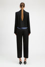 A model shown from the back wearing a black cropped blazer, black cropped trousers, a blue striped shirt, and black heels against a plain white studio background.