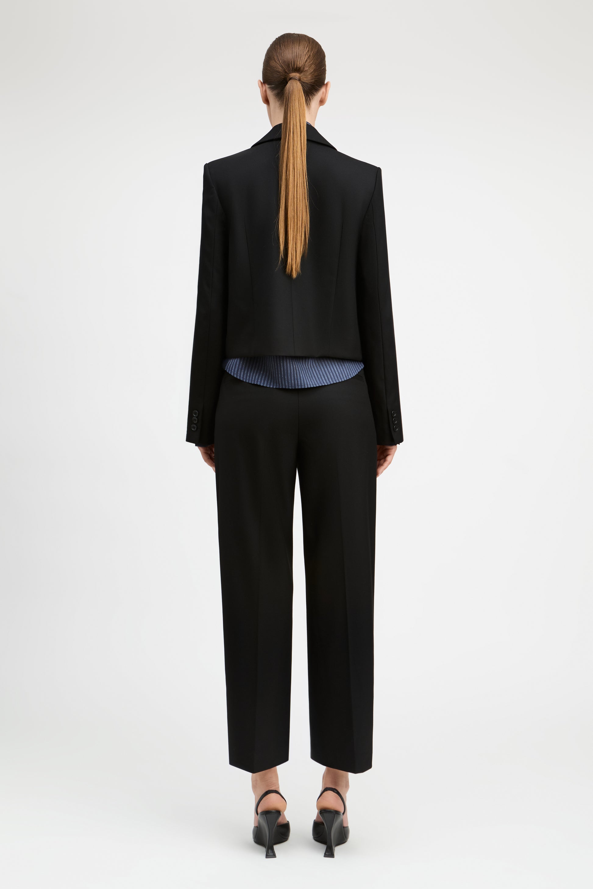 A model shown from the back wearing a black cropped blazer, black cropped trousers, a blue striped shirt, and black heels against a plain white studio background.