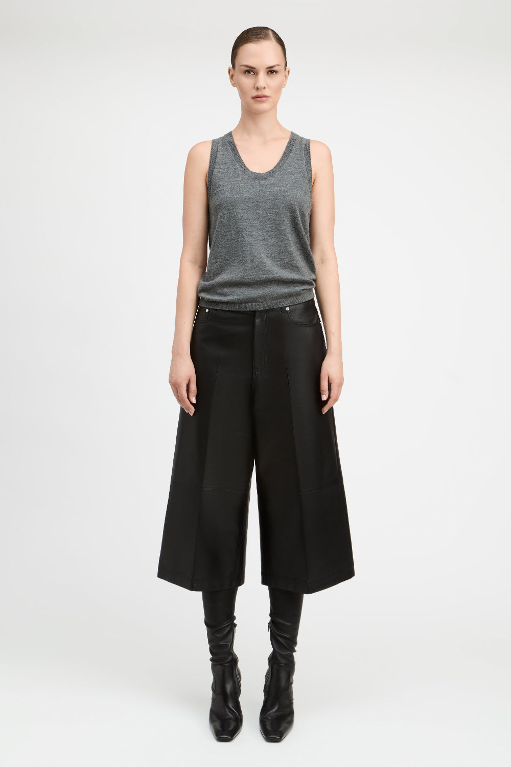 A model stands against a light gray studio background wearing a gray sleeveless knit top, wide-leg black cropped trousers, black leggings, and black pointed boots.