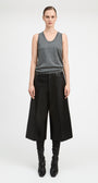 A model stands against a light gray studio background wearing a gray sleeveless knit top, wide-leg black cropped trousers, black leggings, and black pointed boots.