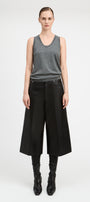 A model stands against a light gray studio background wearing a gray sleeveless knit top, wide-leg black cropped trousers, black leggings, and black pointed boots.