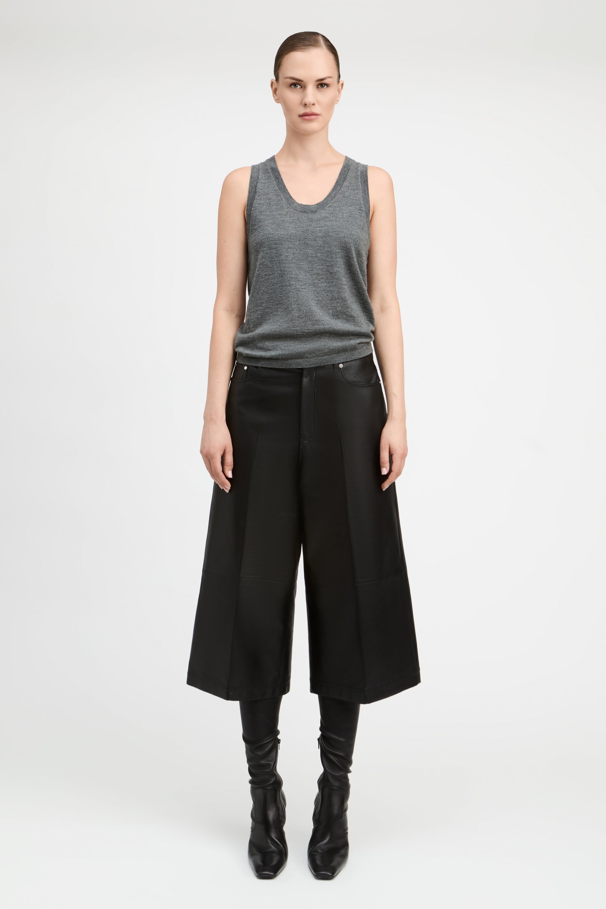A model stands against a light gray studio background wearing a gray sleeveless knit top, wide-leg black cropped trousers, black leggings, and black pointed boots.