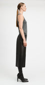 A woman stands in profile wearing a gray sleeveless top, a long black skirt, black leggings, and black heeled boots against a plain light gray studio background.