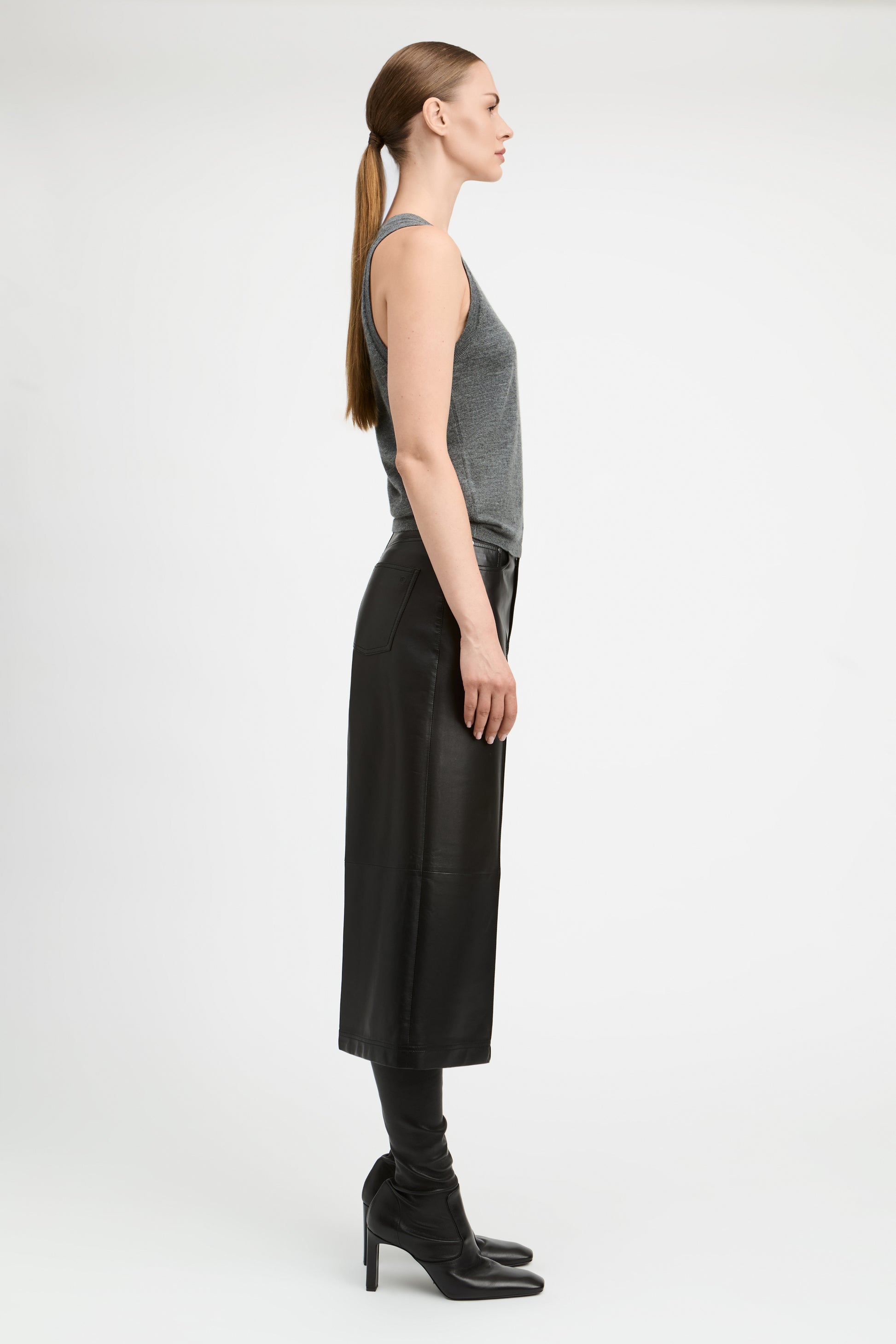 A woman stands in profile wearing a gray sleeveless top, a long black skirt, black leggings, and black heeled boots against a plain light gray studio background.