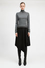 A model stands against a light gray studio background wearing a fitted gray long-sleeve turtleneck sweater, a black asymmetrical draped skirt, black leggings, and black pointed boots.