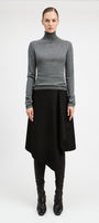 A model stands against a light gray studio background wearing a fitted gray long-sleeve turtleneck sweater, a black asymmetrical draped skirt, black leggings, and black pointed boots.