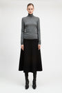 A model stands against a light gray studio background wearing a fitted gray turtleneck sweater, a long black skirt, and black knee-high boots.