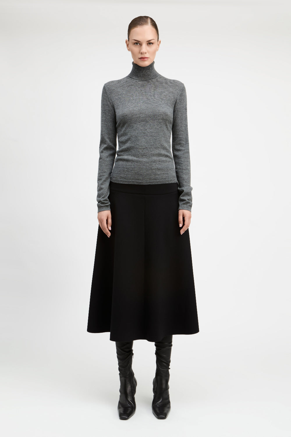 A model stands against a light gray studio background wearing a fitted gray turtleneck sweater, a long black skirt, and black knee-high boots.