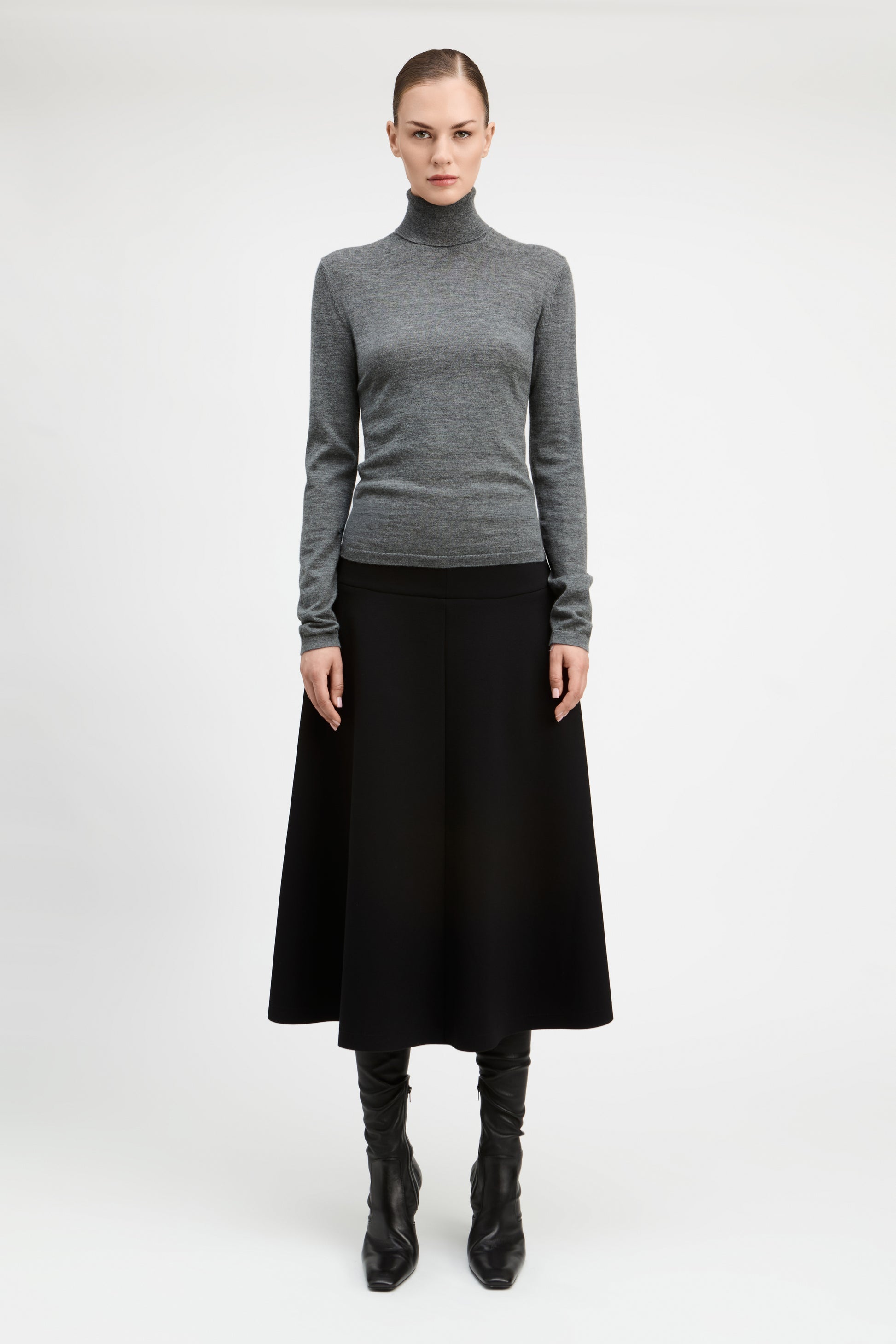 A model stands against a light gray studio background wearing a fitted gray turtleneck sweater, a long black skirt, and black knee-high boots.