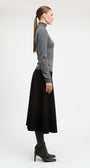 A woman in profile wearing a gray long-sleeve turtleneck sweater, a black midi skirt, black tights, and black heeled ankle boots, photographed against a clean light gray studio background.