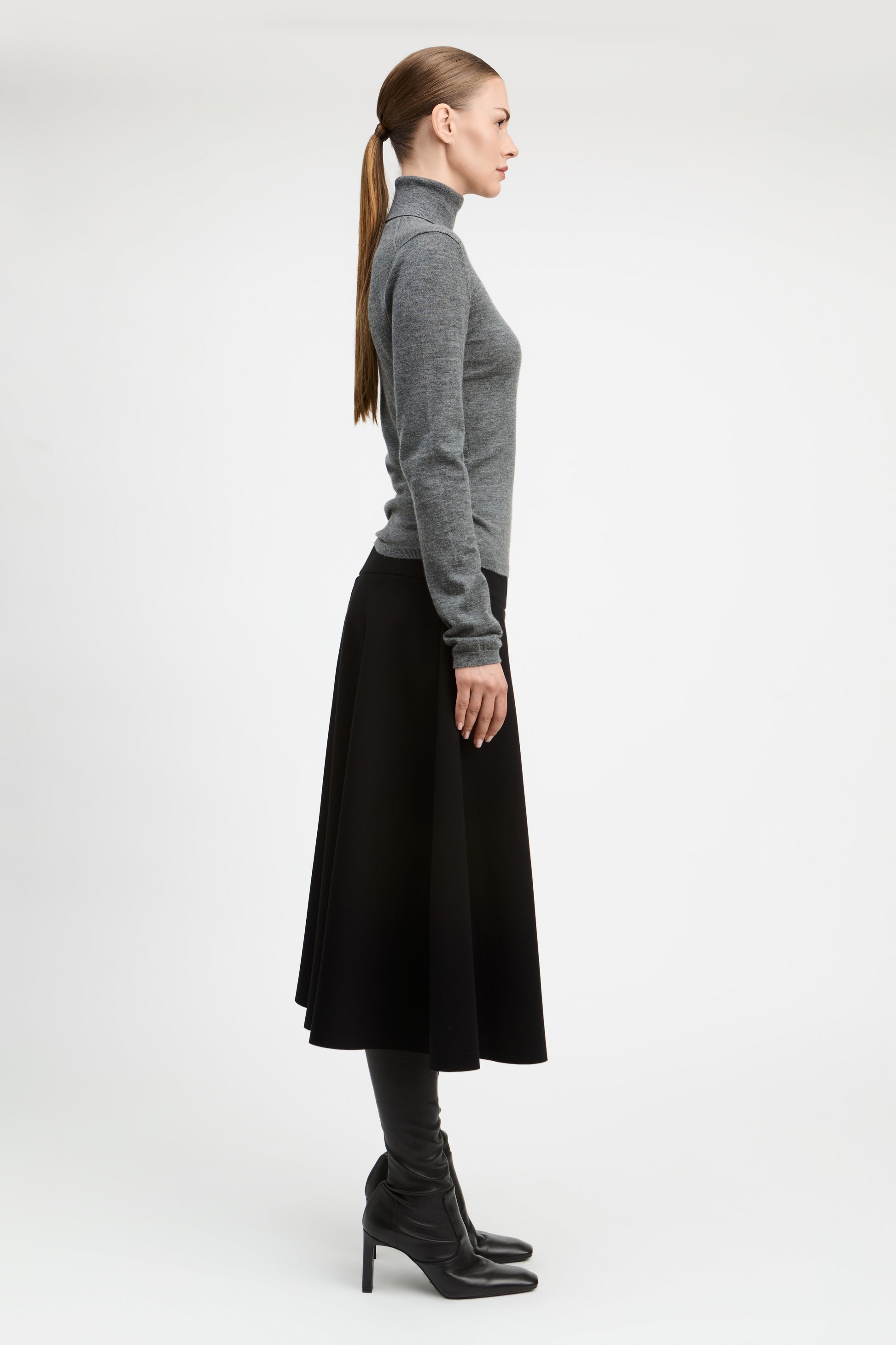 A woman in profile wearing a gray long-sleeve turtleneck sweater, a black midi skirt, black tights, and black heeled ankle boots, photographed against a clean light gray studio background.