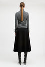 A woman shown from the back wearing a gray long-sleeve mock-neck top, a black midi skirt, black boots, and a brown ponytail, photographed against a plain light gray studio background.