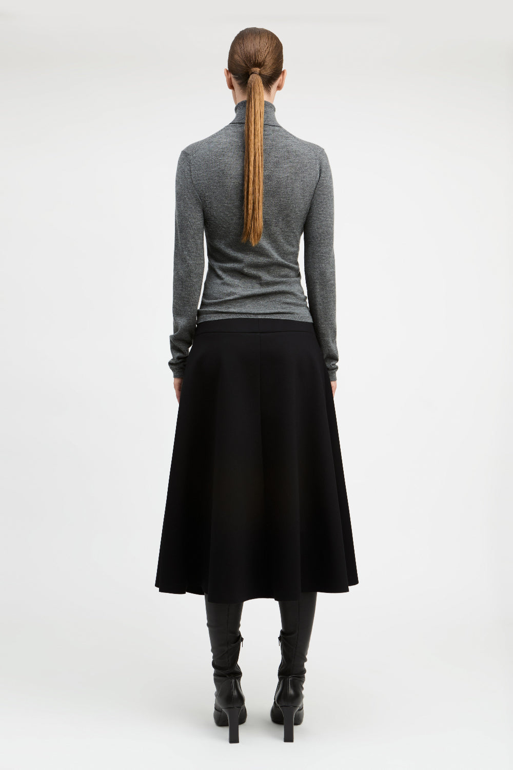 A woman shown from the back wearing a gray long-sleeve mock-neck top, a black midi skirt, black boots, and a brown ponytail, photographed against a plain light gray studio background.