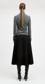 A woman shown from the back wearing a gray long-sleeve mock-neck top, a black midi skirt, black boots, and a brown ponytail, photographed against a plain light gray studio background.