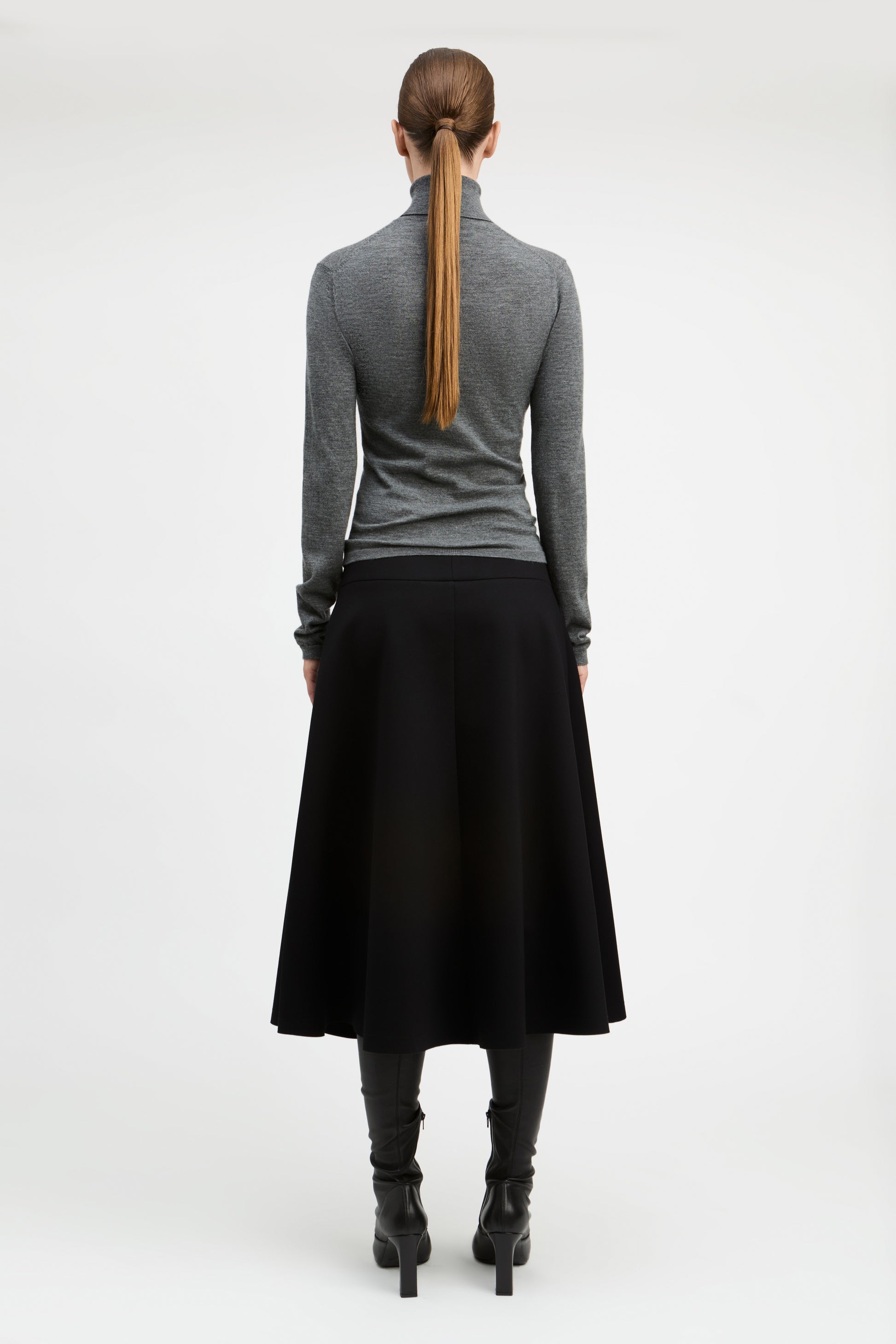 A woman shown from the back wearing a gray long-sleeve mock-neck top, a black midi skirt, black boots, and a brown ponytail, photographed against a plain light gray studio background.