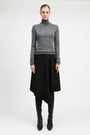 A model stands against a light gray studio background wearing a fitted gray long-sleeve turtleneck sweater, a black asymmetrical draped skirt, black leggings, and black pointed boots.