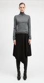 A model stands against a light gray studio background wearing a fitted gray long-sleeve turtleneck sweater, a black asymmetrical draped skirt, black leggings, and black pointed boots.