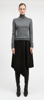 A model stands against a light gray studio background wearing a fitted gray long-sleeve turtleneck sweater, a black asymmetrical draped skirt, black leggings, and black pointed boots.