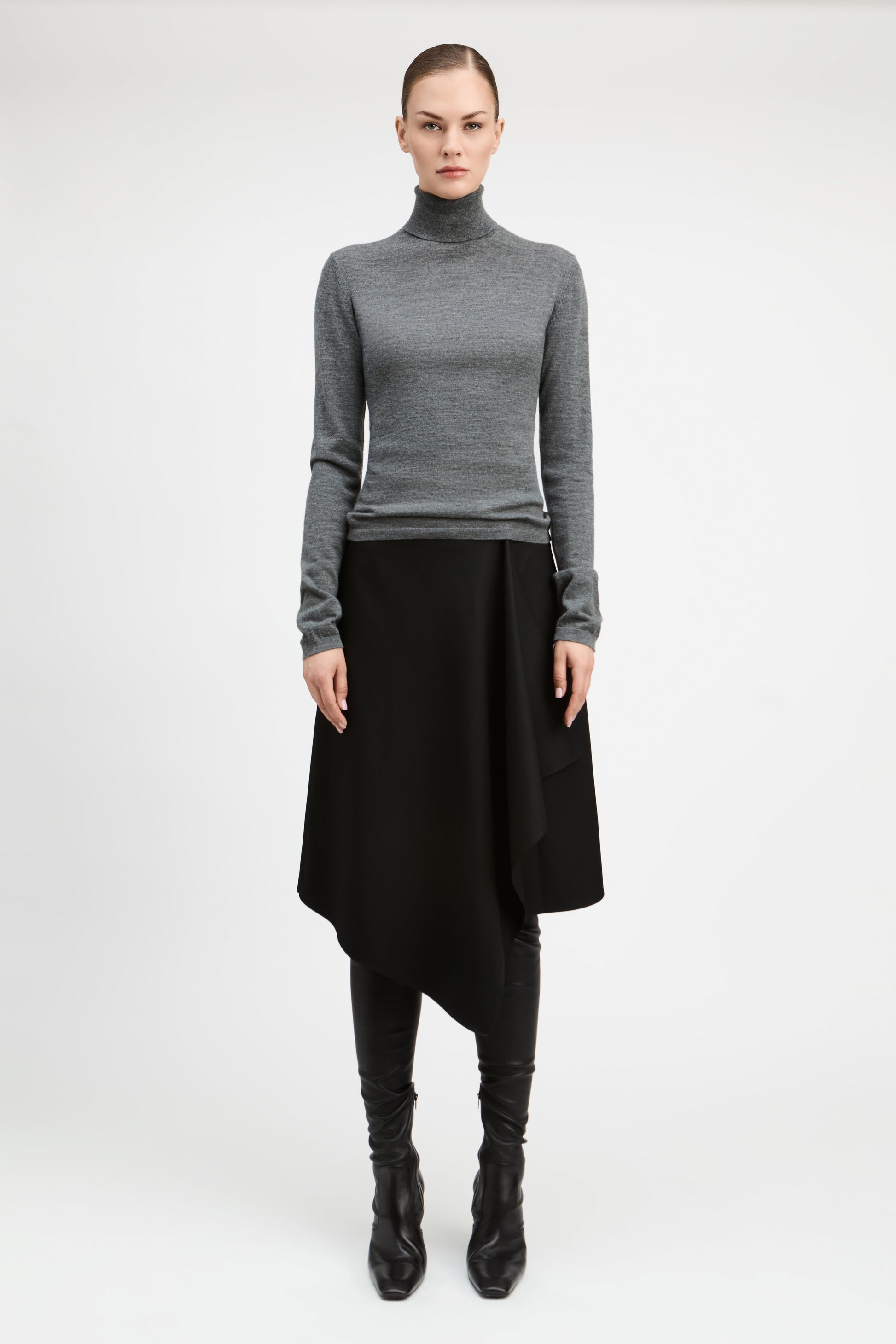 A model stands against a light gray studio background wearing a fitted gray long-sleeve turtleneck sweater, a black asymmetrical draped skirt, black leggings, and black pointed boots.