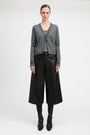 A model stands against a light gray studio background wearing a gray knit cardigan over a dark top, black wide-leg cropped trousers, black boots, and a black belt.