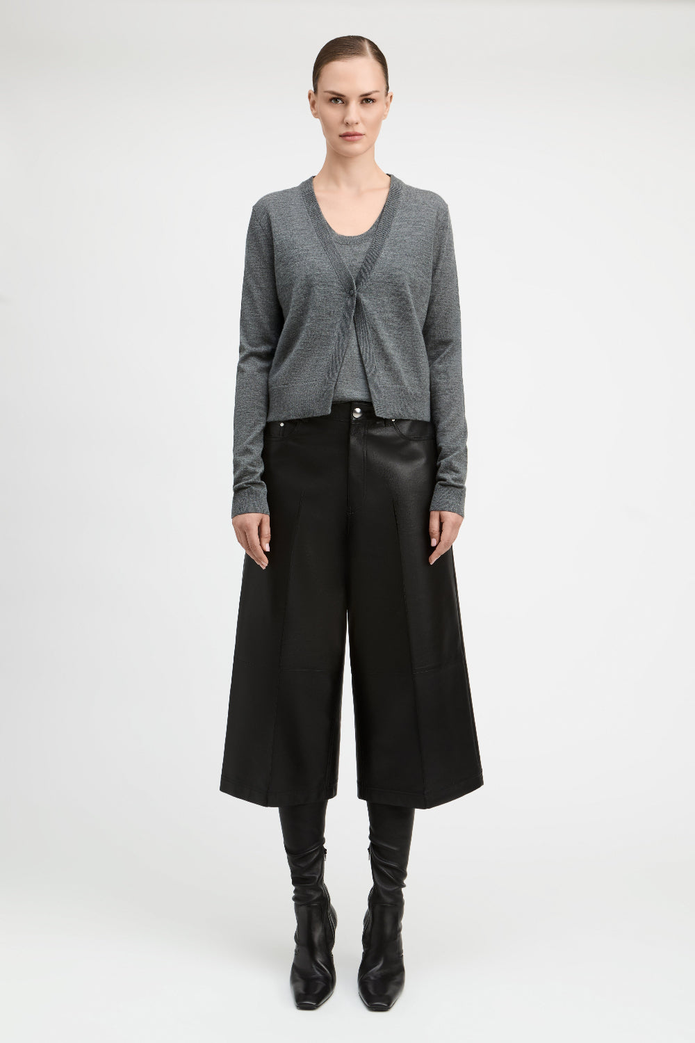 A model stands against a light gray studio background wearing a gray knit cardigan over a dark top, black wide-leg cropped trousers, black boots, and a black belt.