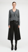 A model stands against a light gray studio background wearing a gray knit cardigan over a dark top, black wide-leg cropped trousers, black boots, and a black belt.