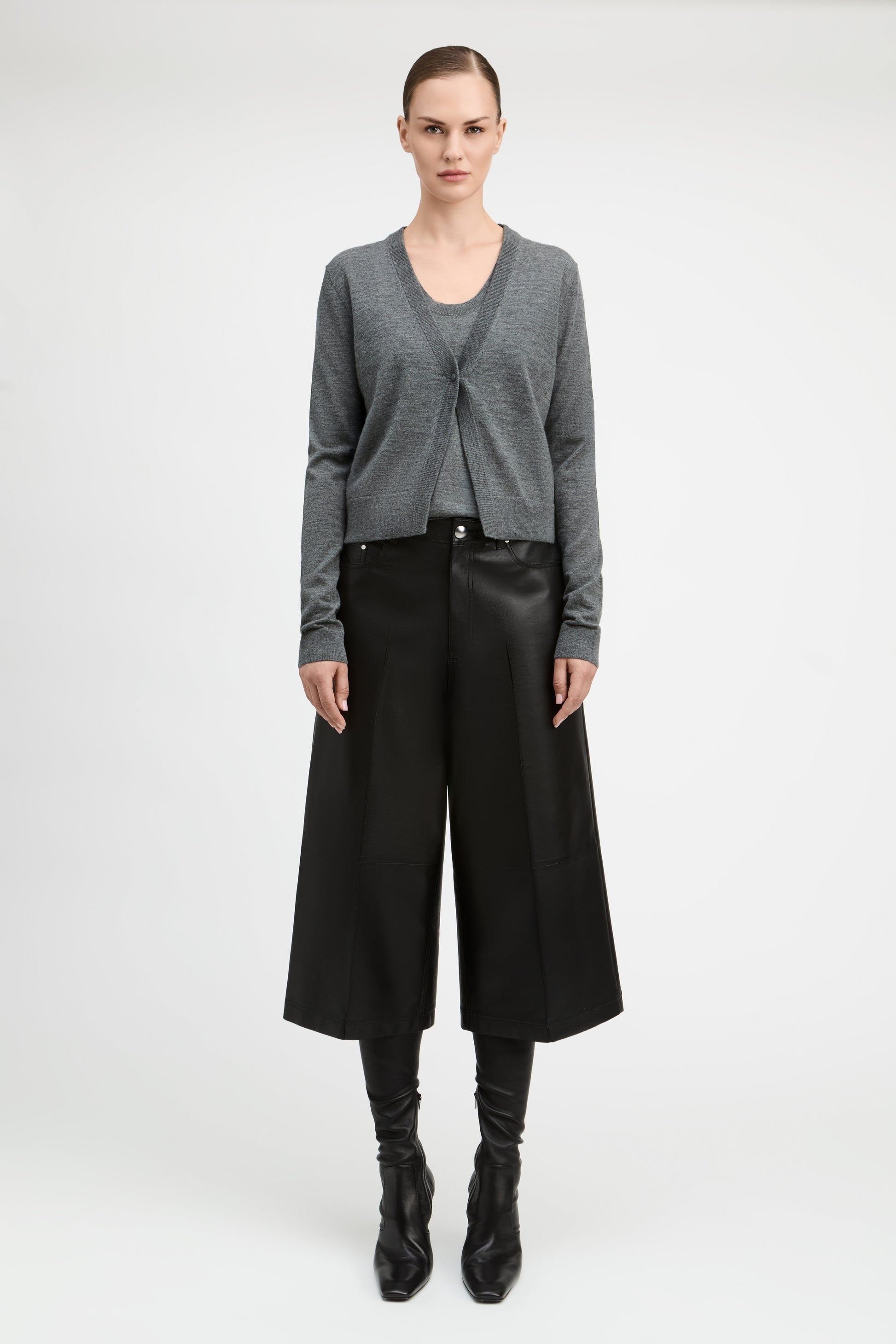 A model stands against a light gray studio background wearing a gray knit cardigan over a dark top, black wide-leg cropped trousers, black boots, and a black belt.