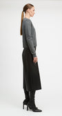 A woman in side profile wearing a gray long-sleeve cropped sweater, a black midi skirt, and black heeled boots, photographed against a clean light gray studio background.