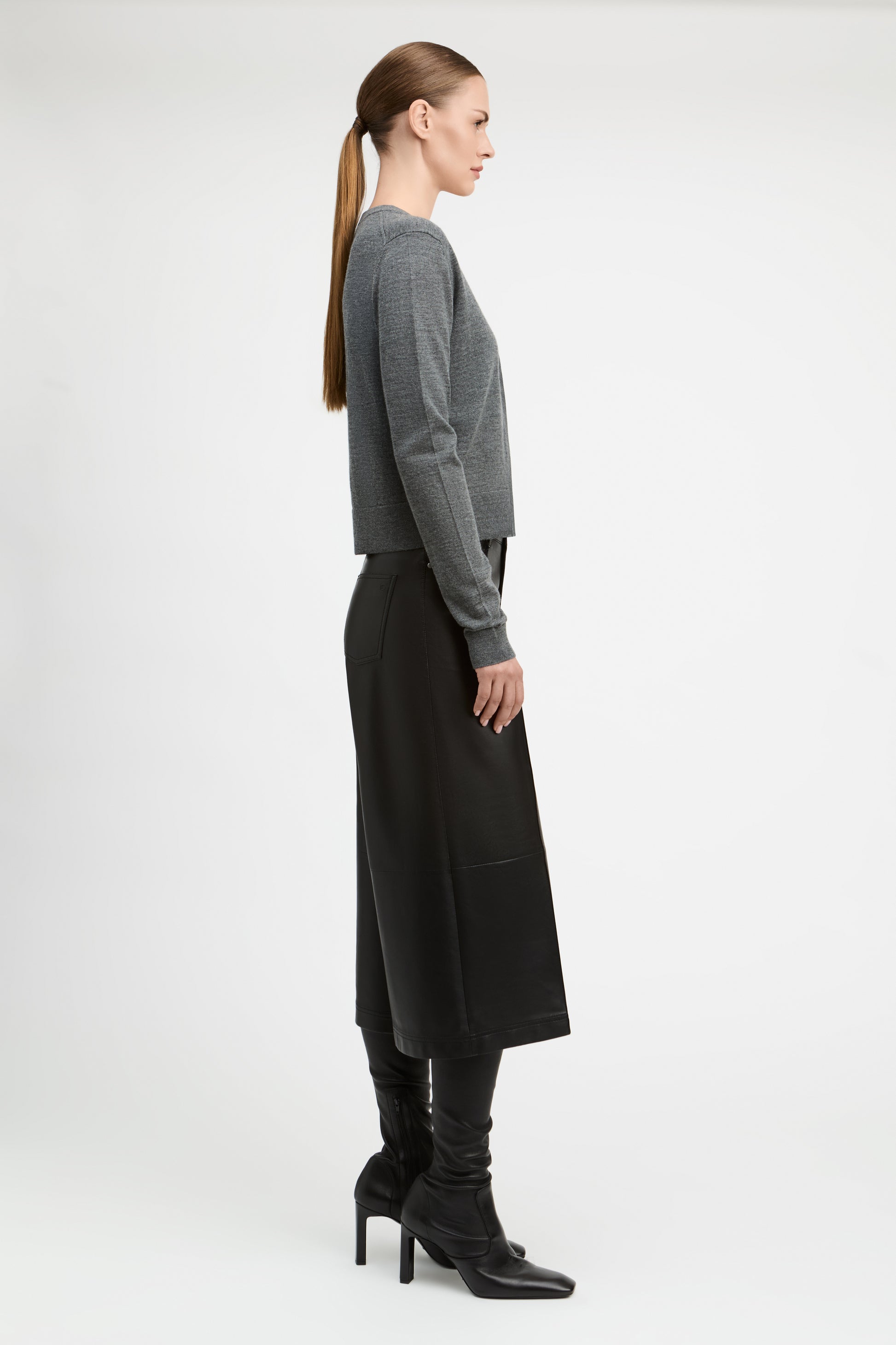 A woman in side profile wearing a gray long-sleeve cropped sweater, a black midi skirt, and black heeled boots, photographed against a clean light gray studio background.