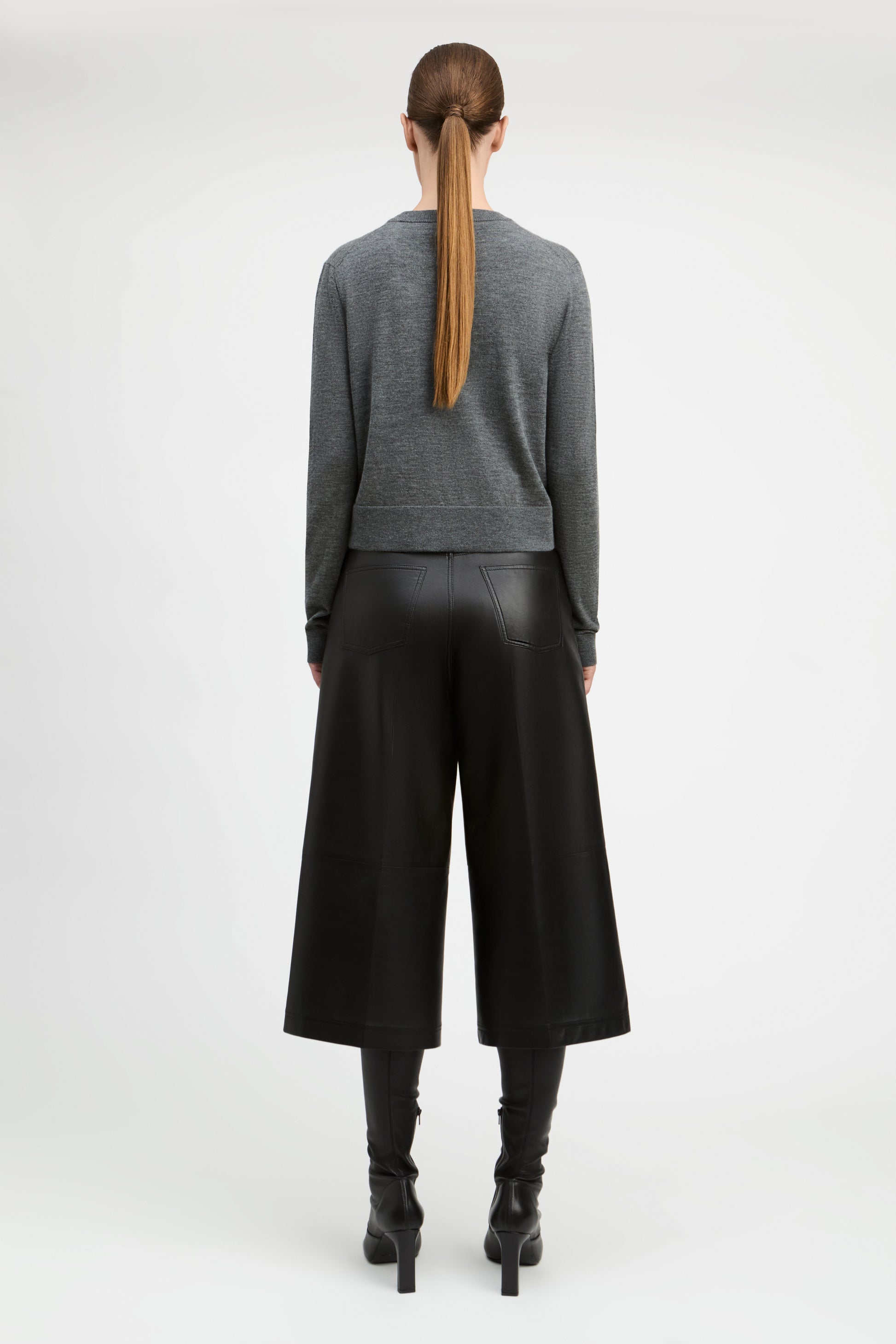 A model viewed from the back wearing a gray long-sleeve knit sweater, black wide-leg cropped pants, and black heeled boots against a plain light gray studio background.