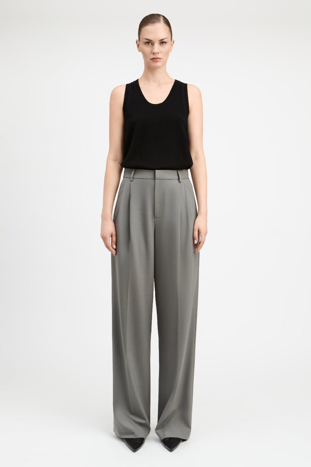 A model stands against a light gray studio background wearing a black sleeveless top and high-waisted, wide-leg gray trousers.