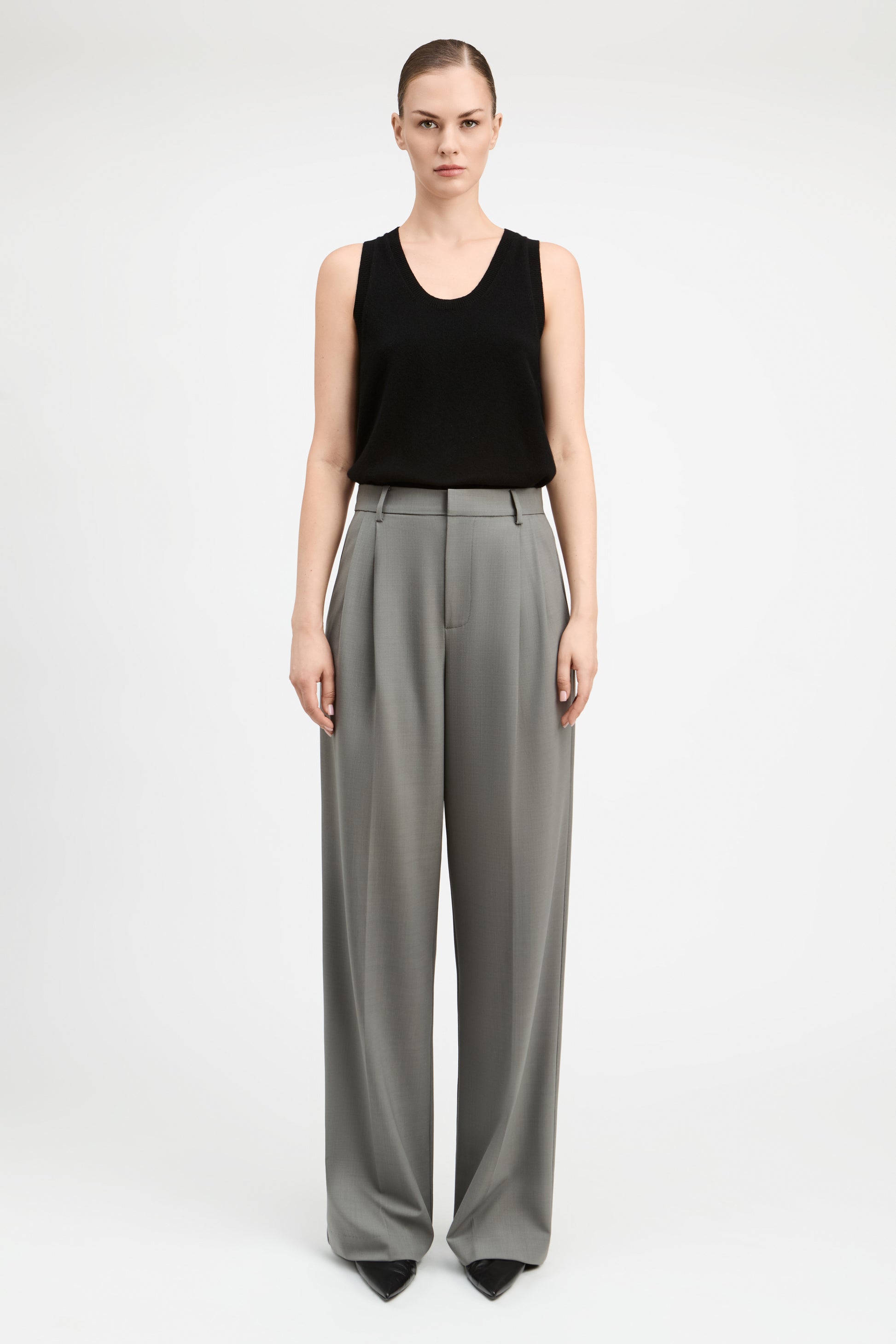 A model stands against a light gray studio background wearing a black sleeveless top and high-waisted, wide-leg gray trousers.