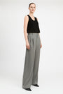 A model stands against a light gray studio background wearing a black sleeveless top and high-waisted, wide-leg gray trousers with black pointed shoes.