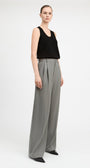A model stands against a light gray studio background wearing a black sleeveless top and high-waisted, wide-leg gray trousers with black pointed shoes.