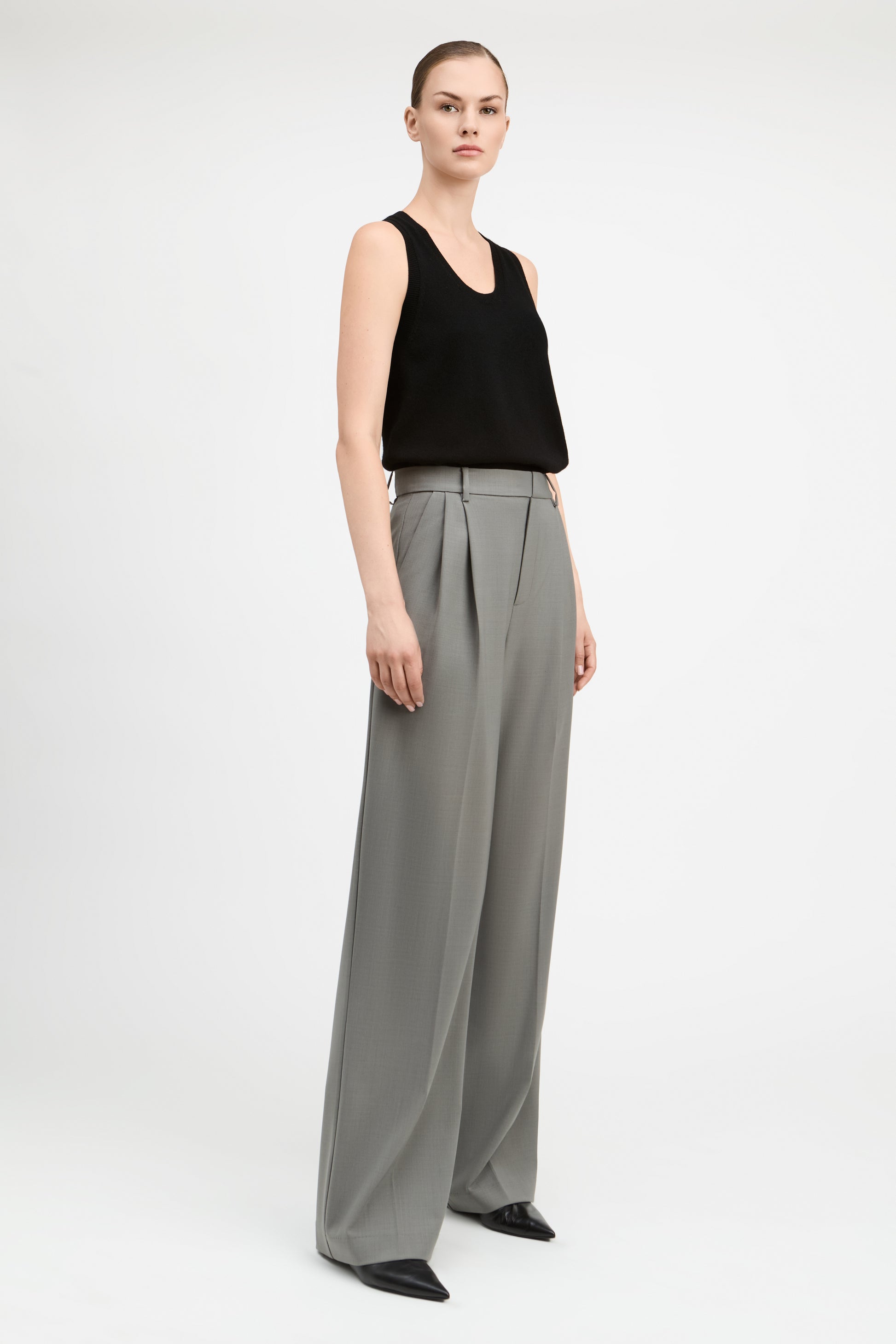 A model stands against a light gray studio background wearing a black sleeveless top and high-waisted, wide-leg gray trousers with black pointed shoes.