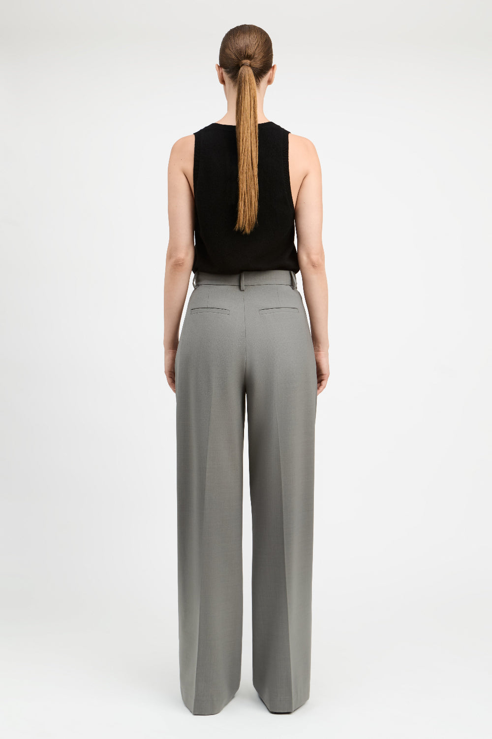 A woman shown from the back wearing a black sleeveless top and high-waisted, wide-leg gray trousers against a plain white studio background.