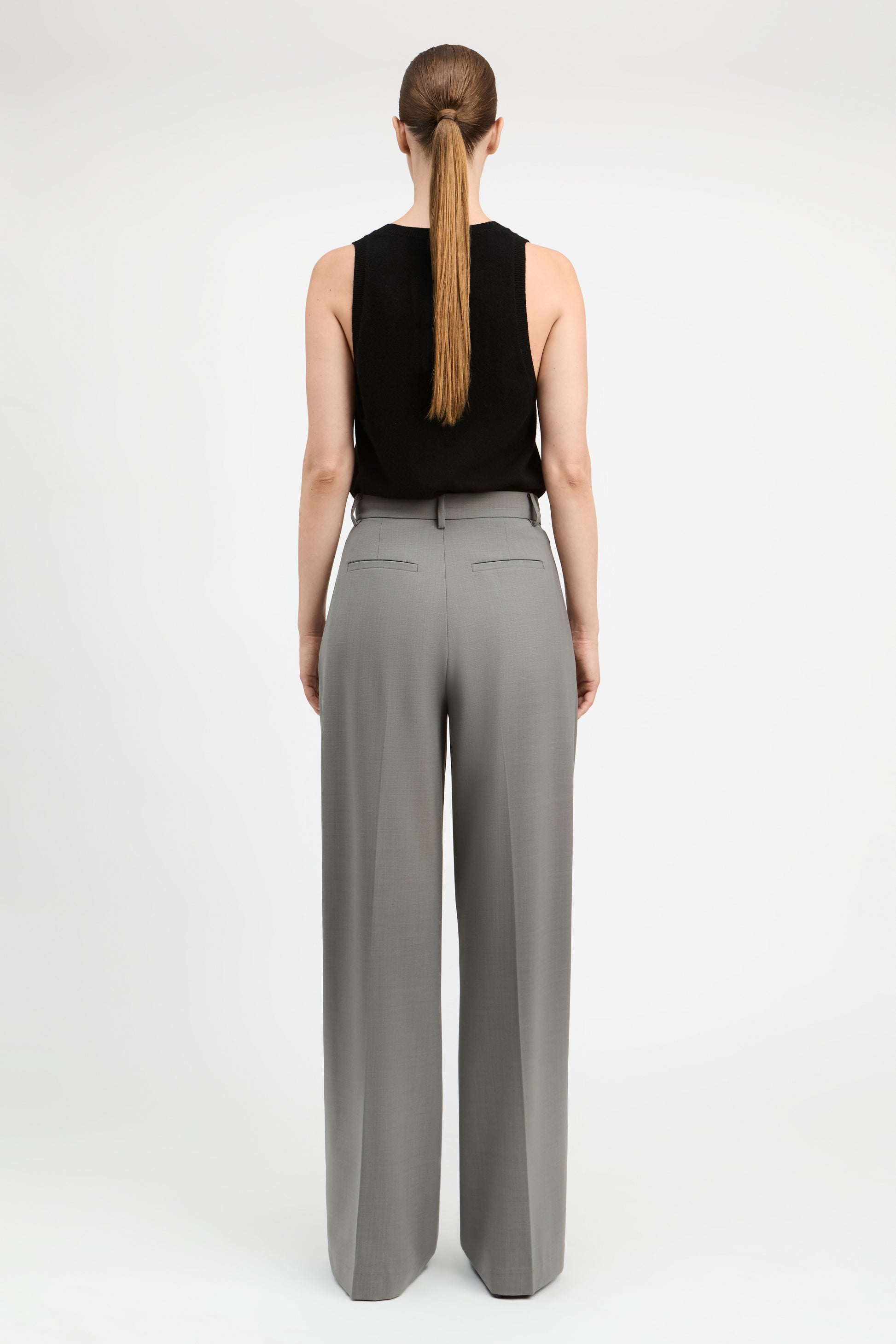 A woman shown from the back wearing a black sleeveless top and high-waisted, wide-leg gray trousers against a plain white studio background.