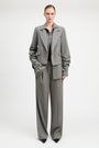A model stands against a light gray studio background wearing a tailored gray suit with a structured blazer and matching wide-leg trousers, paired with a dark top and black pointed shoes.