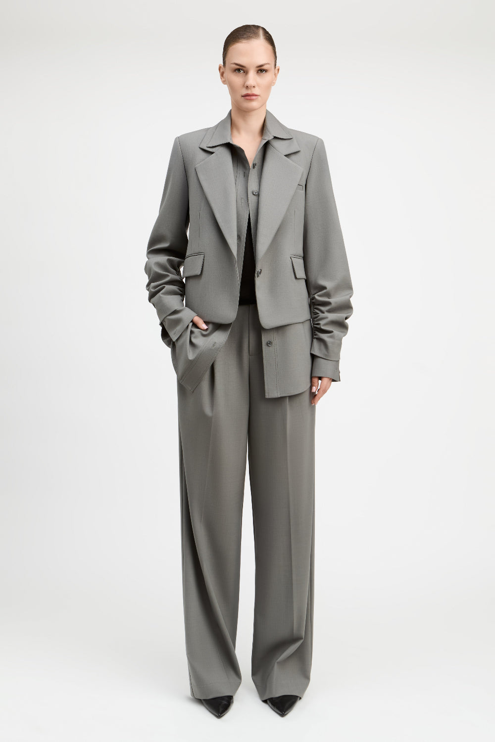 A model stands against a light gray studio background wearing a tailored gray suit with a structured blazer and matching wide-leg trousers, paired with a dark top and black pointed shoes.