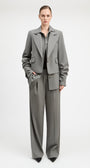 A model stands against a light gray studio background wearing a tailored gray suit with a structured blazer and matching wide-leg trousers, paired with a dark top and black pointed shoes.
